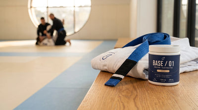 Base / 01 beside a white gi and blue jiujitsu belt with a black rank bar in a sunlit academy